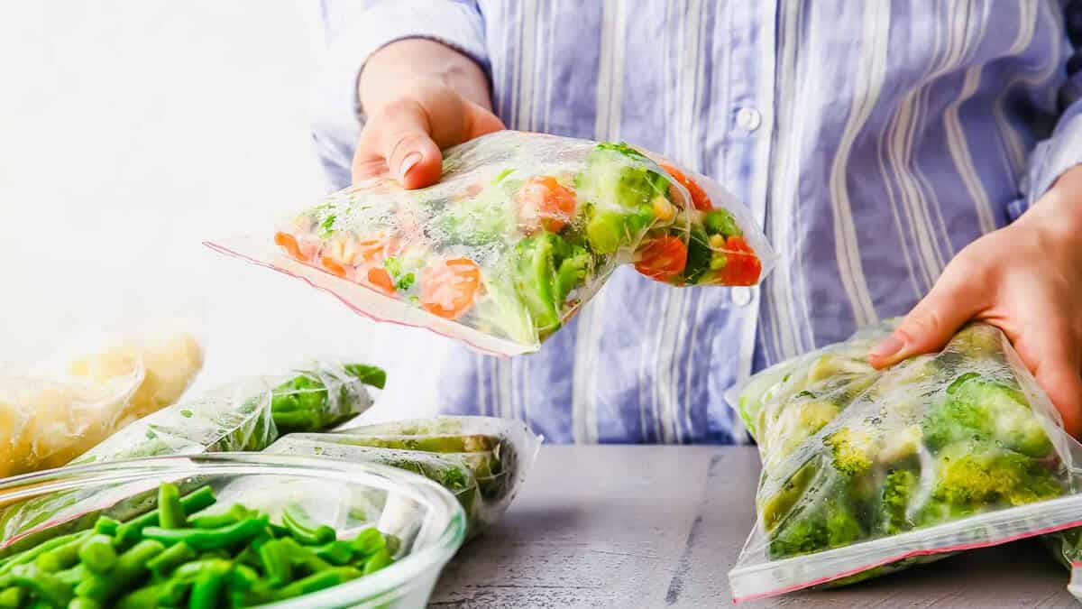 Person places mixed vegetables into a plastic freezer bag for storage, with other bags and vegetables visible on the table.