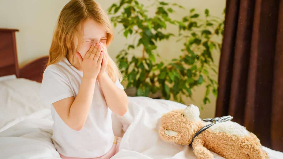 Young girl sitting on a bed, covering her face with hands, crying next to a teddy bear with a stethoscope.