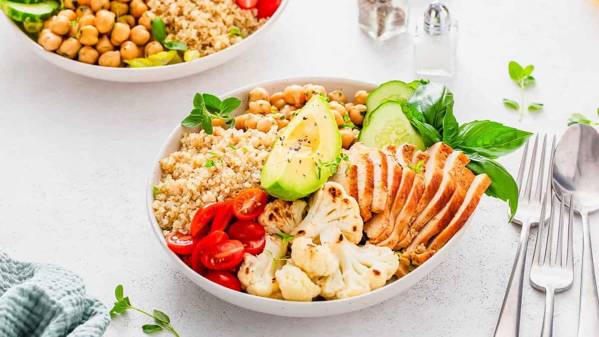 Healthy meal in a bowl with grilled chicken, quinoa, chickpeas, avocado, tomatoes, and cauliflower, garnished with fresh basil.