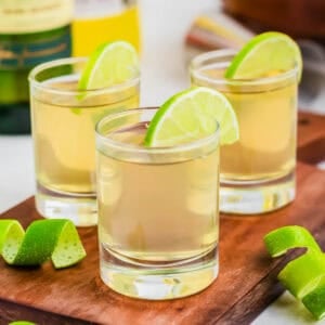 Three green tea shot glasses garnished with lime wedges on wooden cutting board.