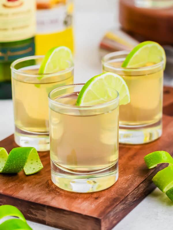 Three green tea shot glasses garnished with lime wedges on wooden cutting board.