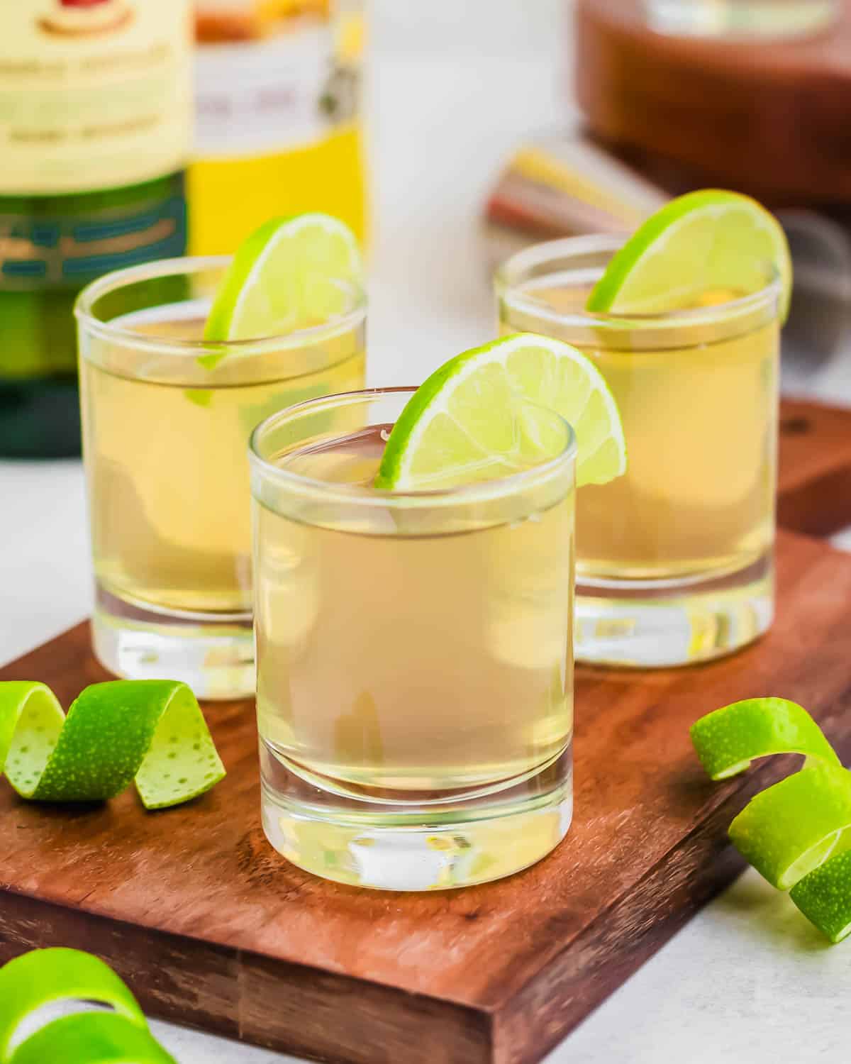 Three green tea shot glasses garnished with lime wedges on wooden cutting board.