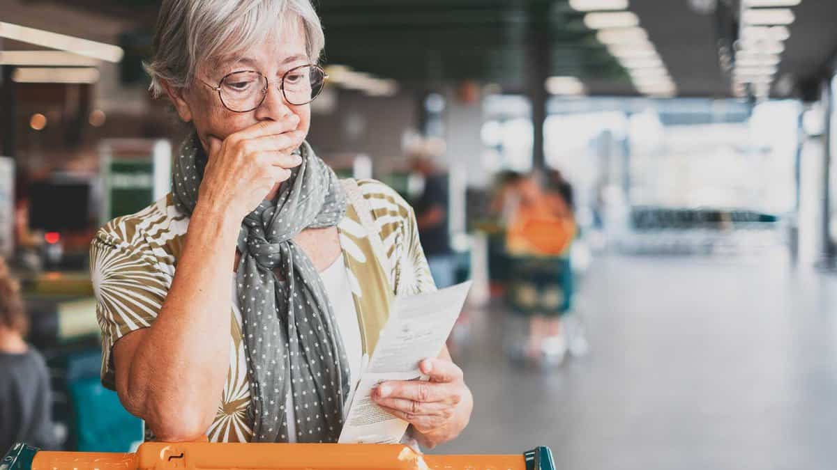 An elderly woman with gray hair and glasses stands in a supermarket, reading a receipt with a thoughtful expression. She is dressed in casual clothing and stands near a shopping cart.