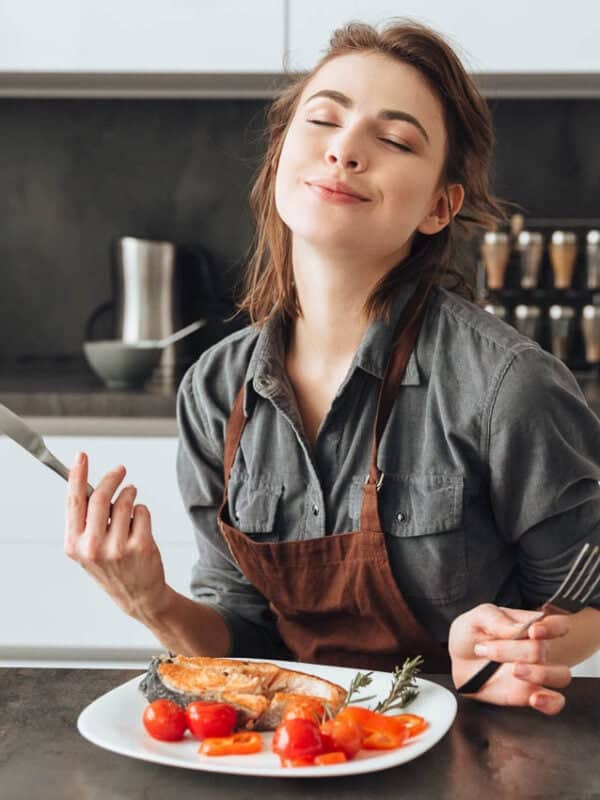 A contented woman in an apron sitting at a kitchen counter, enjoying the aroma of a grilled meal on a plate in front of her.