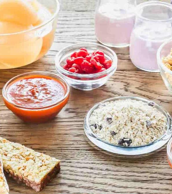 A variety of breakfast items are displayed, including granola bars, dried fruit, granola, yogurt, and fruit sauces in bowls, all arranged on a wooden table.