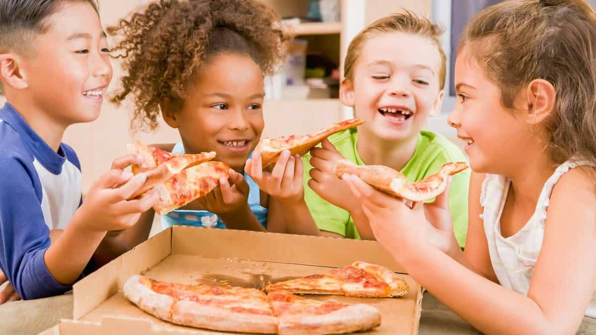 Four children happily eating pizza around a table with an open pizza box in front of them.