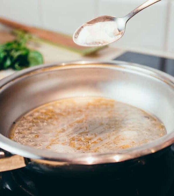 Hand pouring sauce from a spoon into a frying pan over a stove, with chopped vegetables and a bowl nearby in a kitchen.