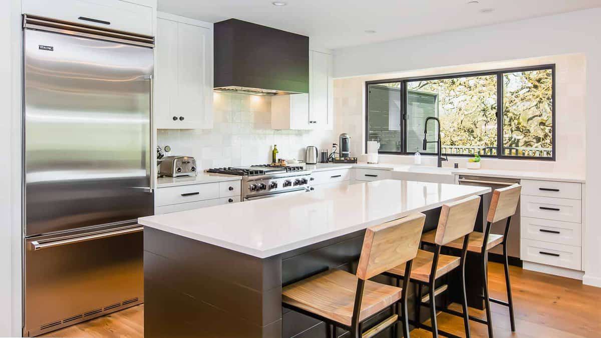 Modern kitchen with stainless steel appliances, white cabinetry, and a large island with three wooden bar stools. A window above the sink offers natural light.