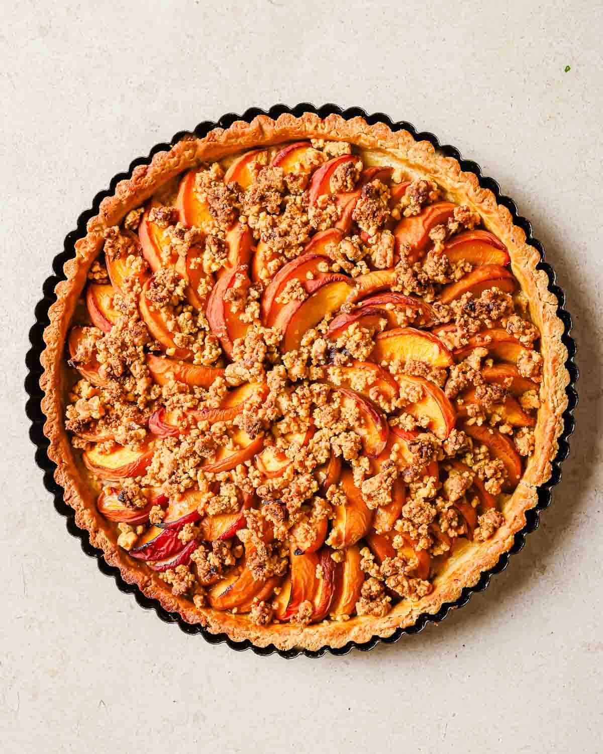 A freshly baked peach tart in a fluted tart pan, topped with sliced peaches and crumble, on a light beige surface.