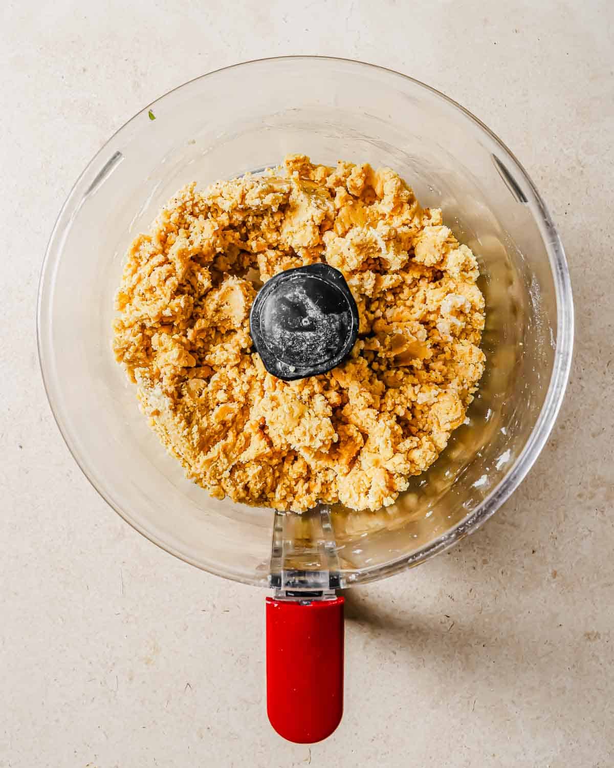 A glass food processor bowl with a red handle, containing a mixture of crumbled dough on a light surface.