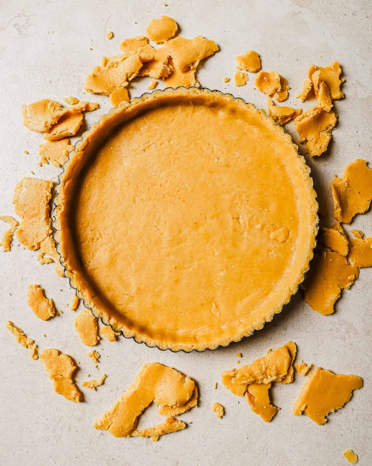 Unbaked tart crust pressed into a tart pan surrounded by scattered crust pieces on a light beige surface.