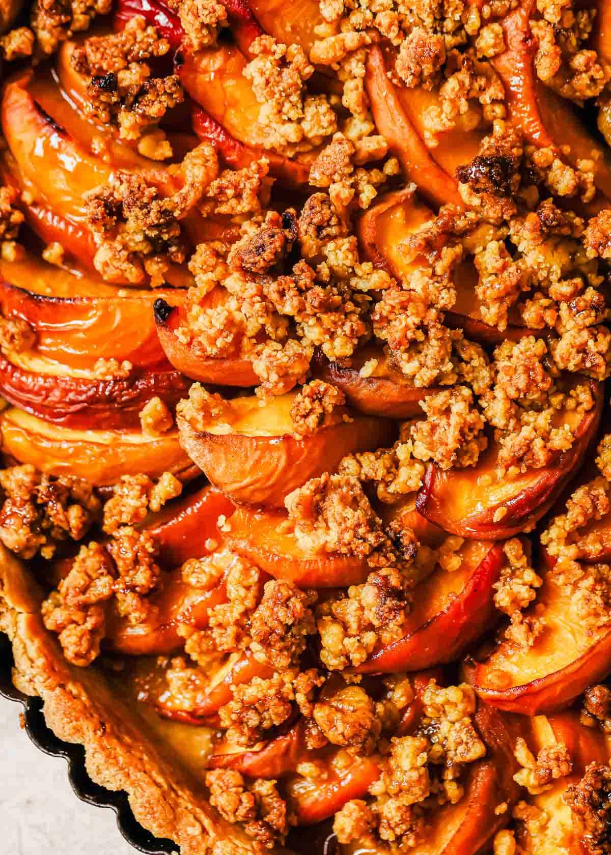 Close-up image of a tart with sliced peaches and crumbled topping, baked to a golden brown, in a black tart pan.