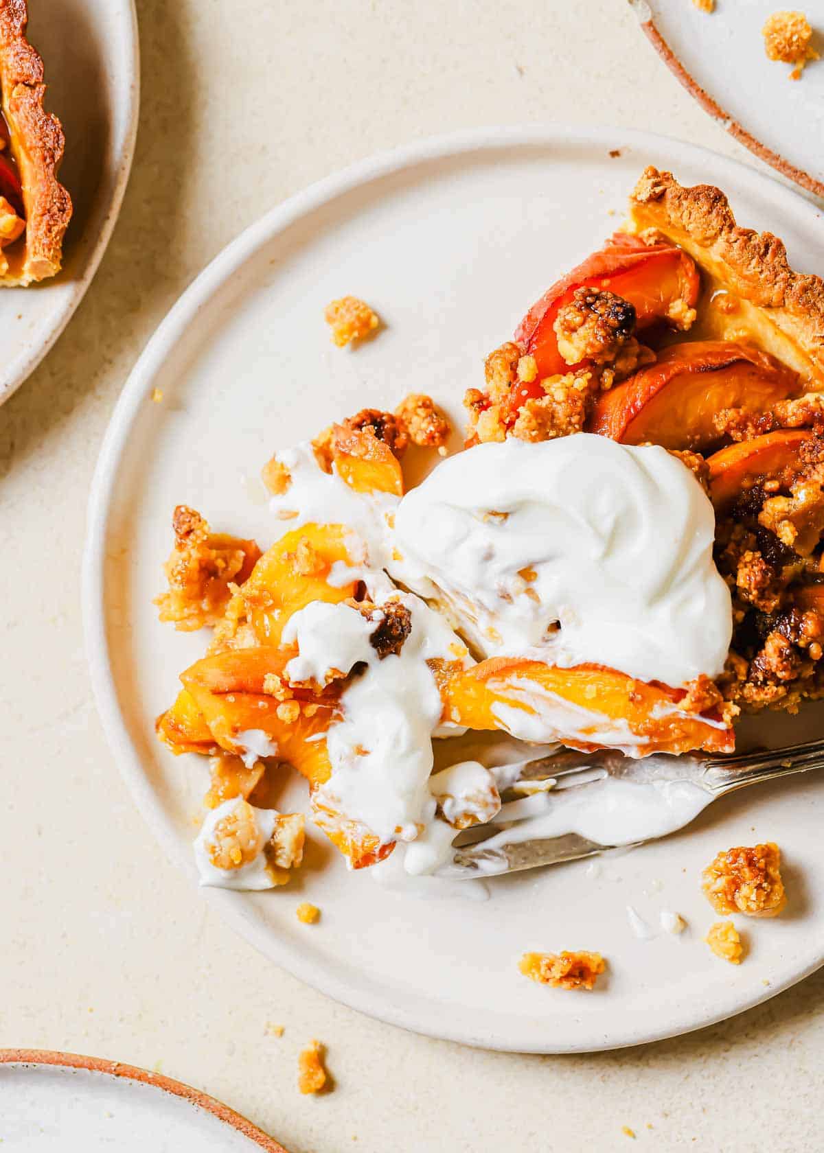 A slice of peach tart topped with whipped cream on a white plate, with a fork taking a bite out of it.