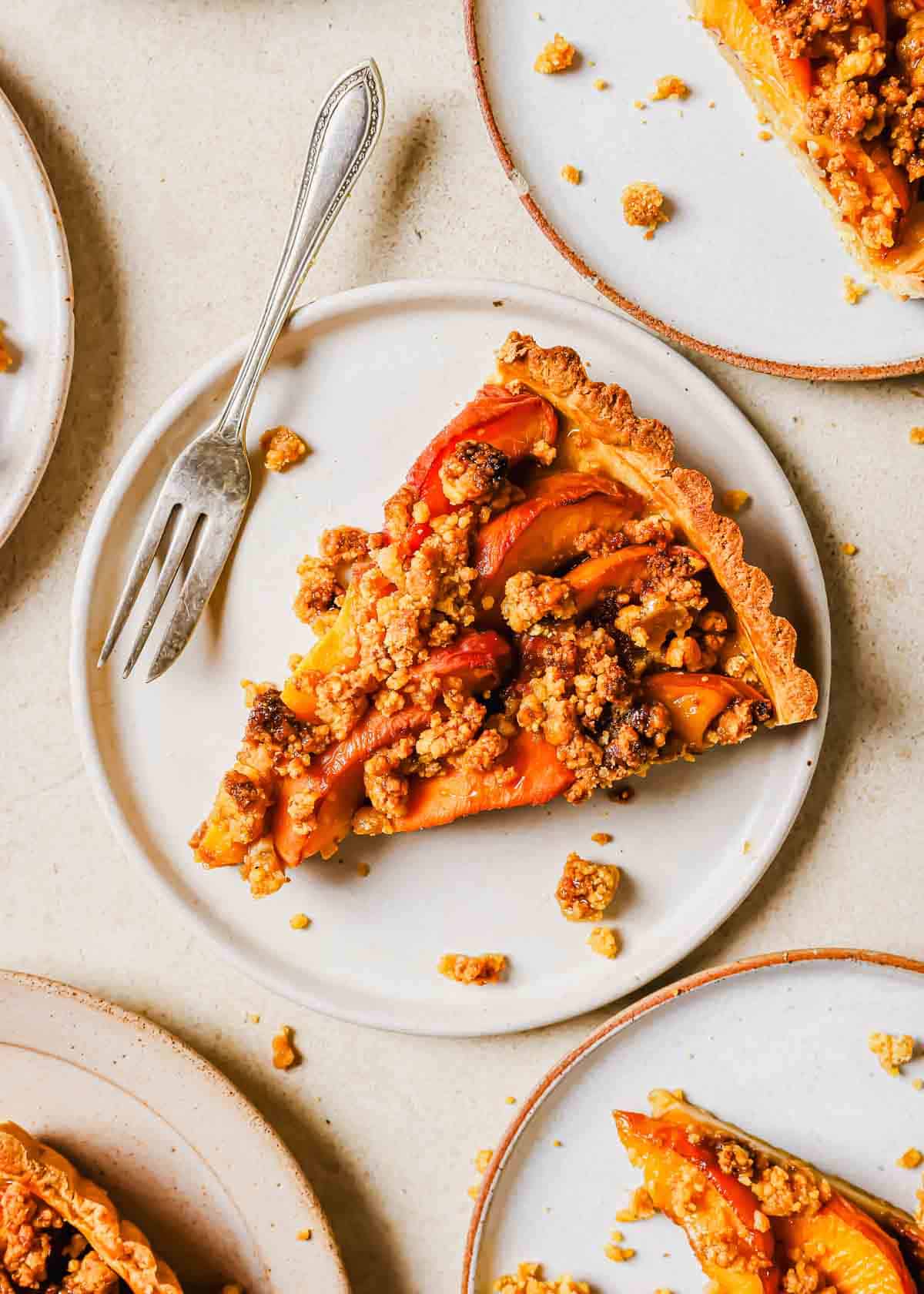 Slices of peach pie with crumb topping on white plates, each served with a fork, on a light speckled surface.