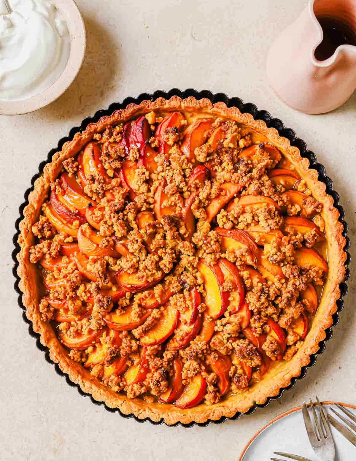 A freshly baked peach tart with sliced peaches and crumble topping, served alongside a pitcher of cream on a beige surface.