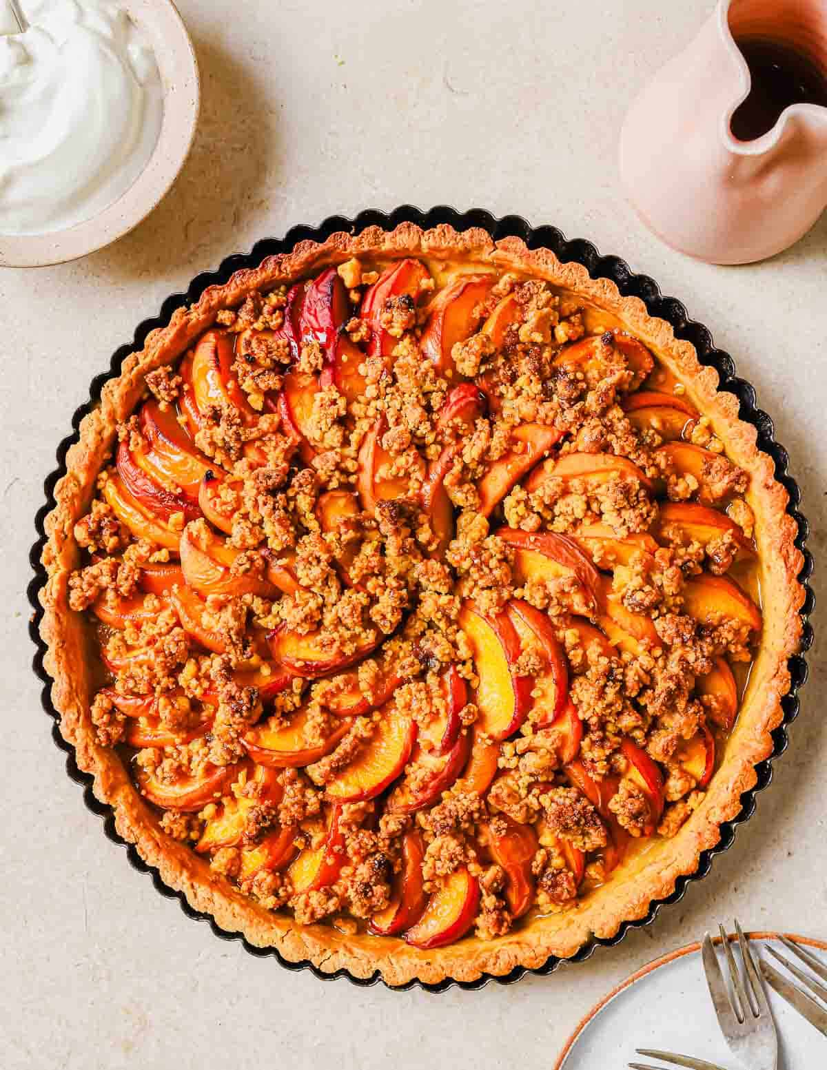 A freshly baked peach tart with sliced peaches and crumble topping, served alongside a pitcher of cream on a beige surface.