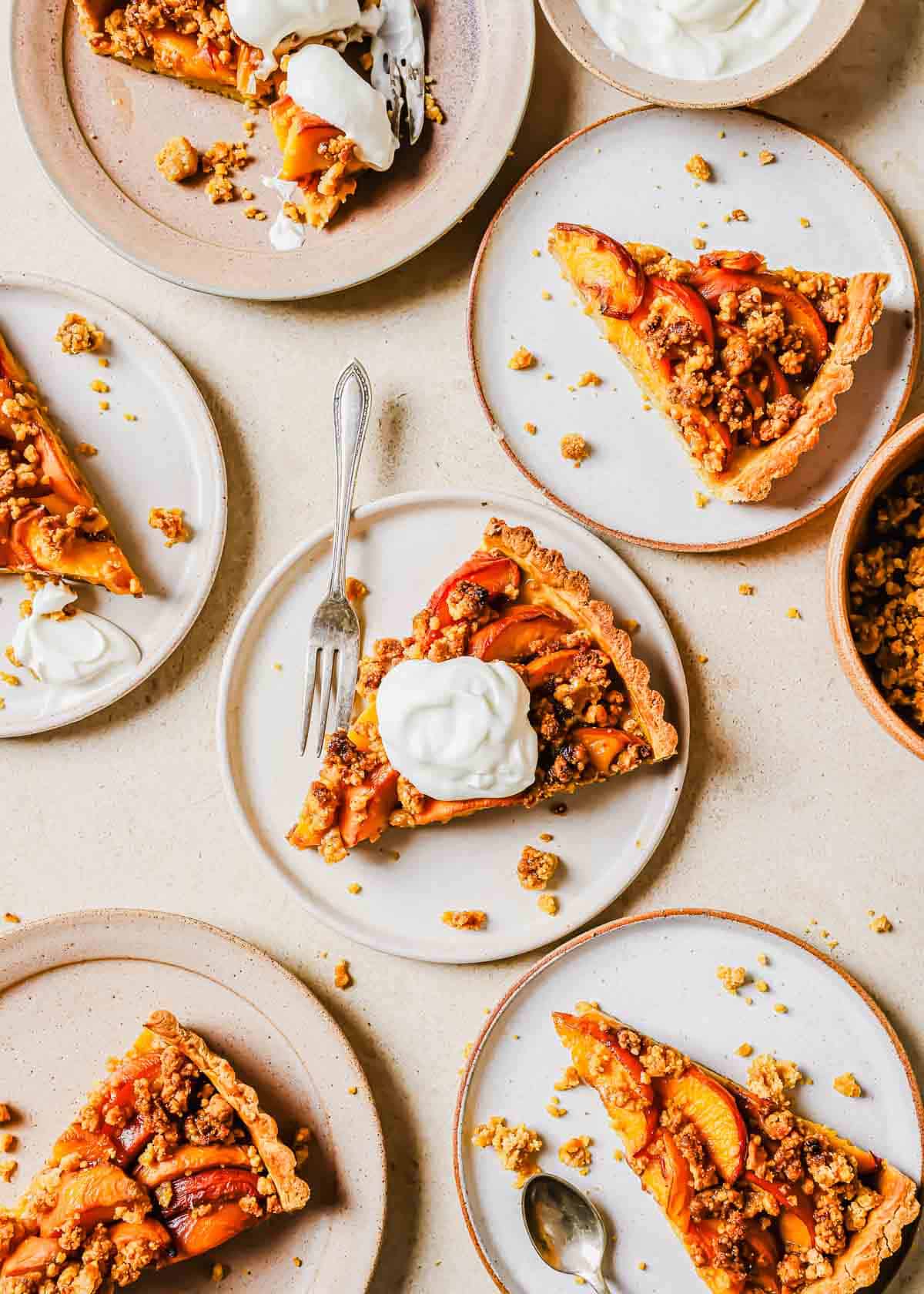 Several plates with slices of peach tart topped with whipped cream on a light surface.