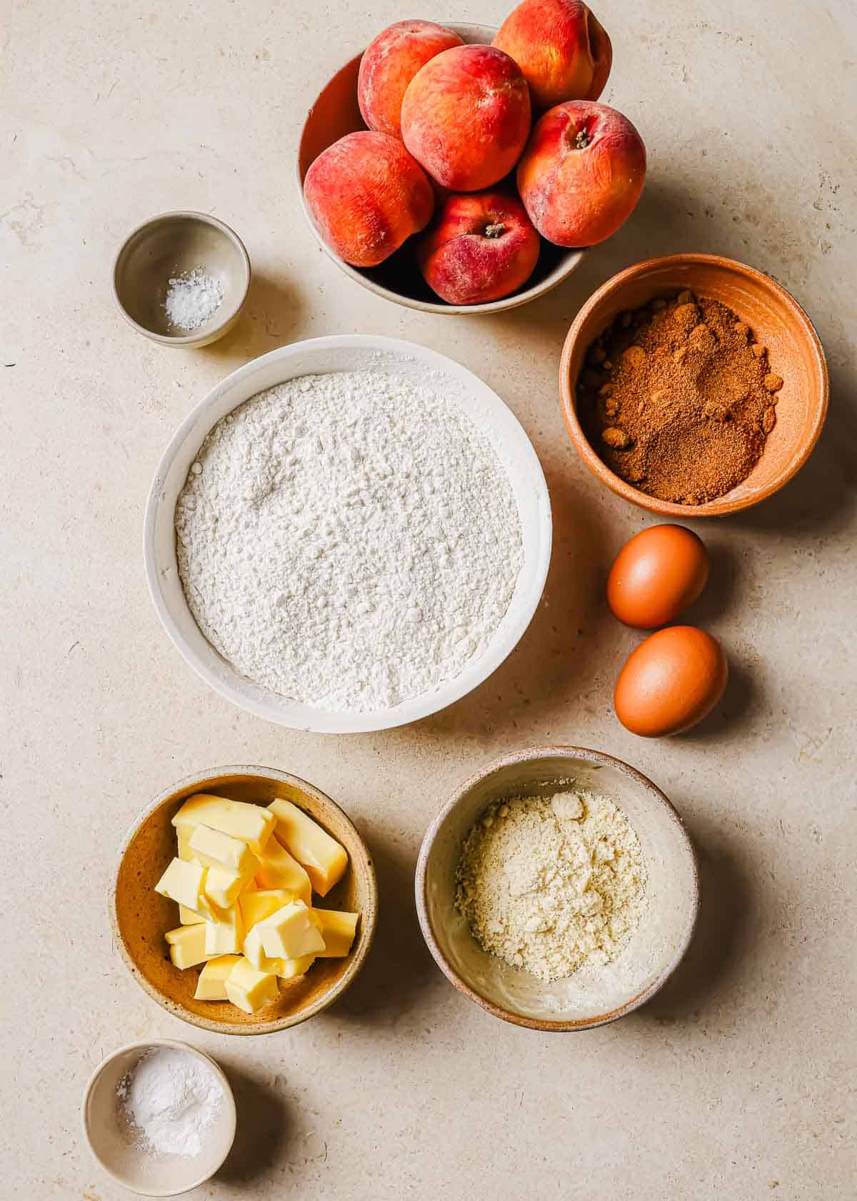 Ingredients for baking a peach tart arranged on a table, including flour, eggs, butter, sugar, peaches, and spices in separate bowls.