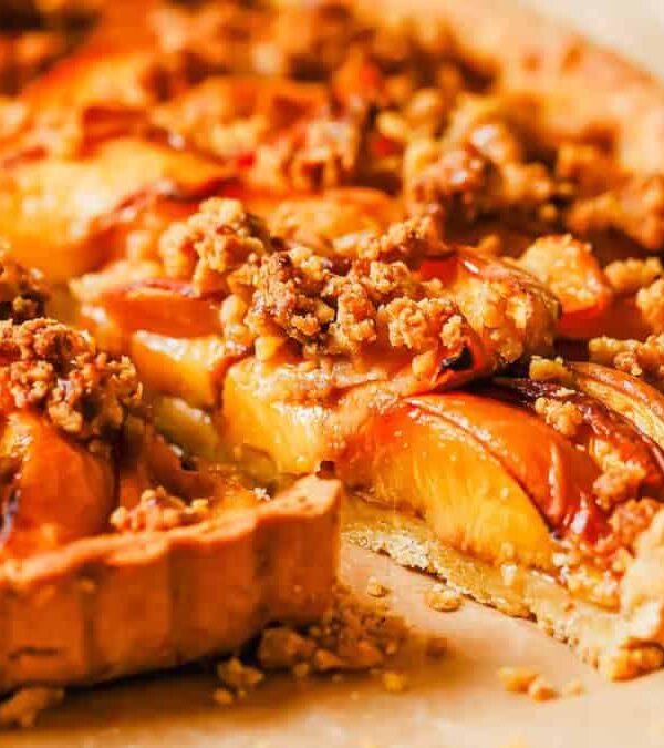 A close-up of a sliced fruit tart topped with crumbled nuts and glazed peaches on a crumbly crust.