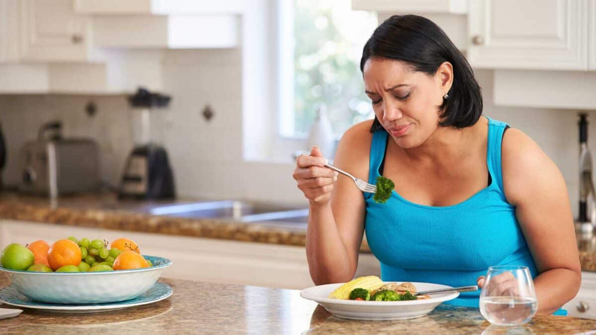 A woman in a blue tank top sits in a kitchen, looking displeased while holding a fork with broccoli. A plate of diverse fruits and a glass of water are on the kitchen counter.