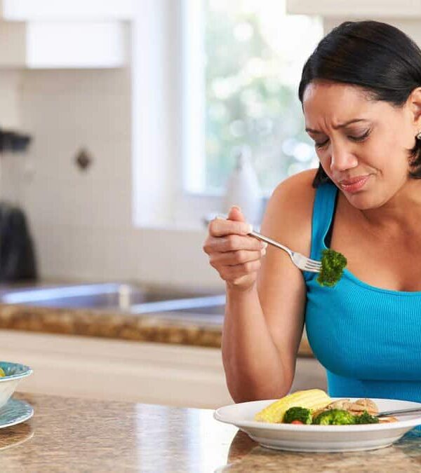 A woman in a blue tank top sits in a kitchen, looking displeased while holding a fork with broccoli. A plate of diverse fruits and a glass of water are on the kitchen counter.