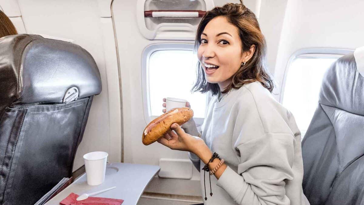 A woman on an airplane seated by the window holds a sandwich and a cup, appearing to smile at the camera.