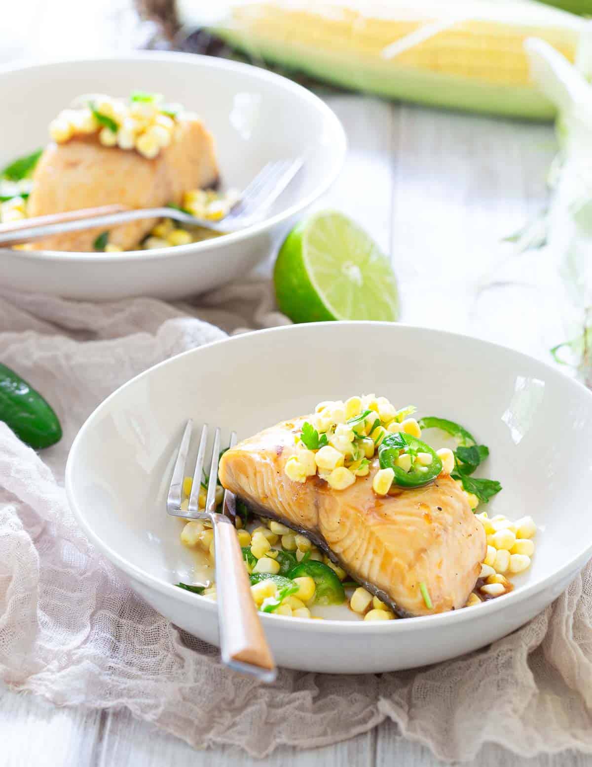 Two white bowls contain cooked sablefish fillets topped with corn and green garnish, with forks resting in the bowls. A lime and corn on the cob are visible in the background.