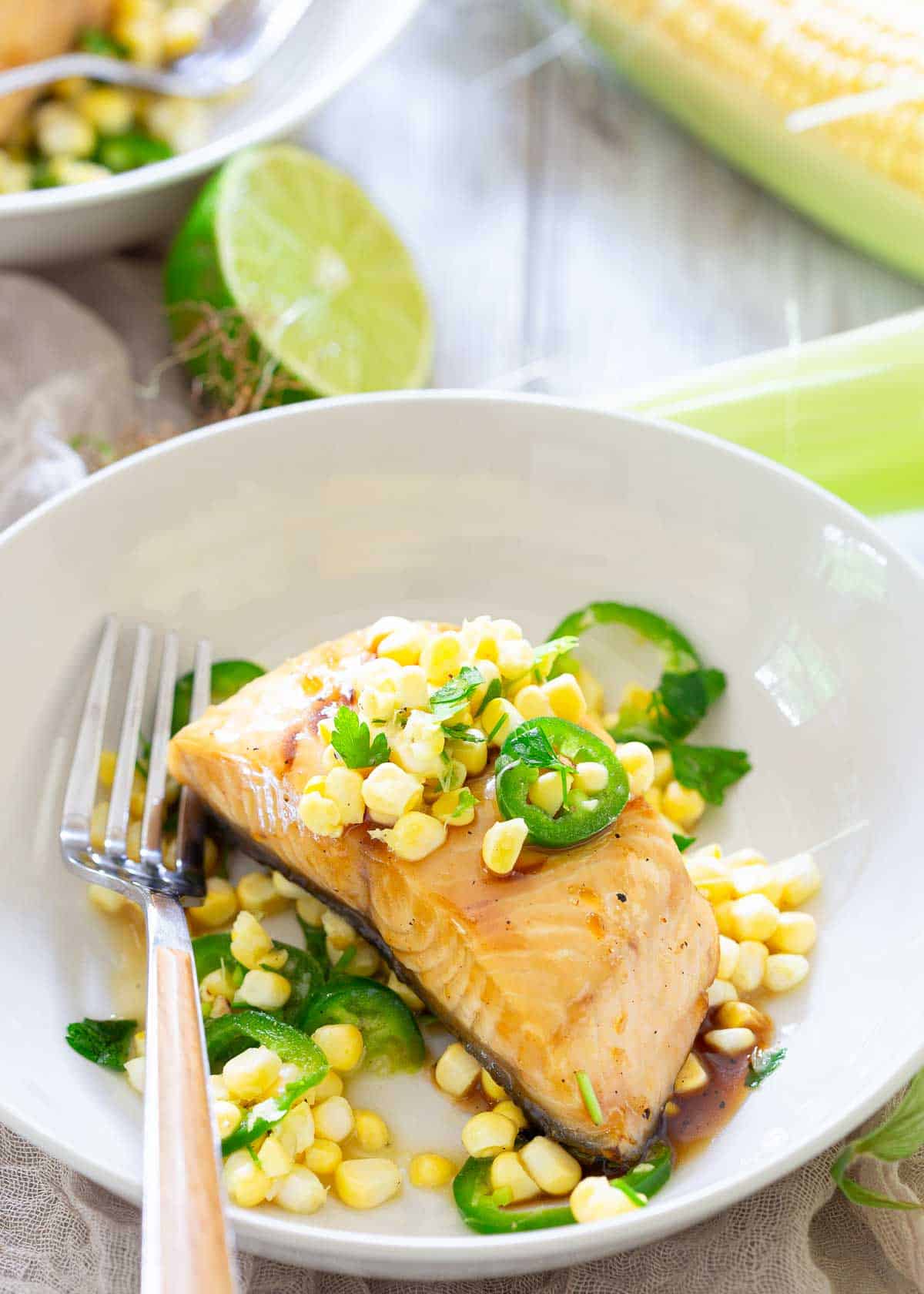 A white plate with a piece of cooked black cod topped with corn kernels and sliced jalapeños, garnished with herbs, next to a fork. A lime and a corn cob are in the background.