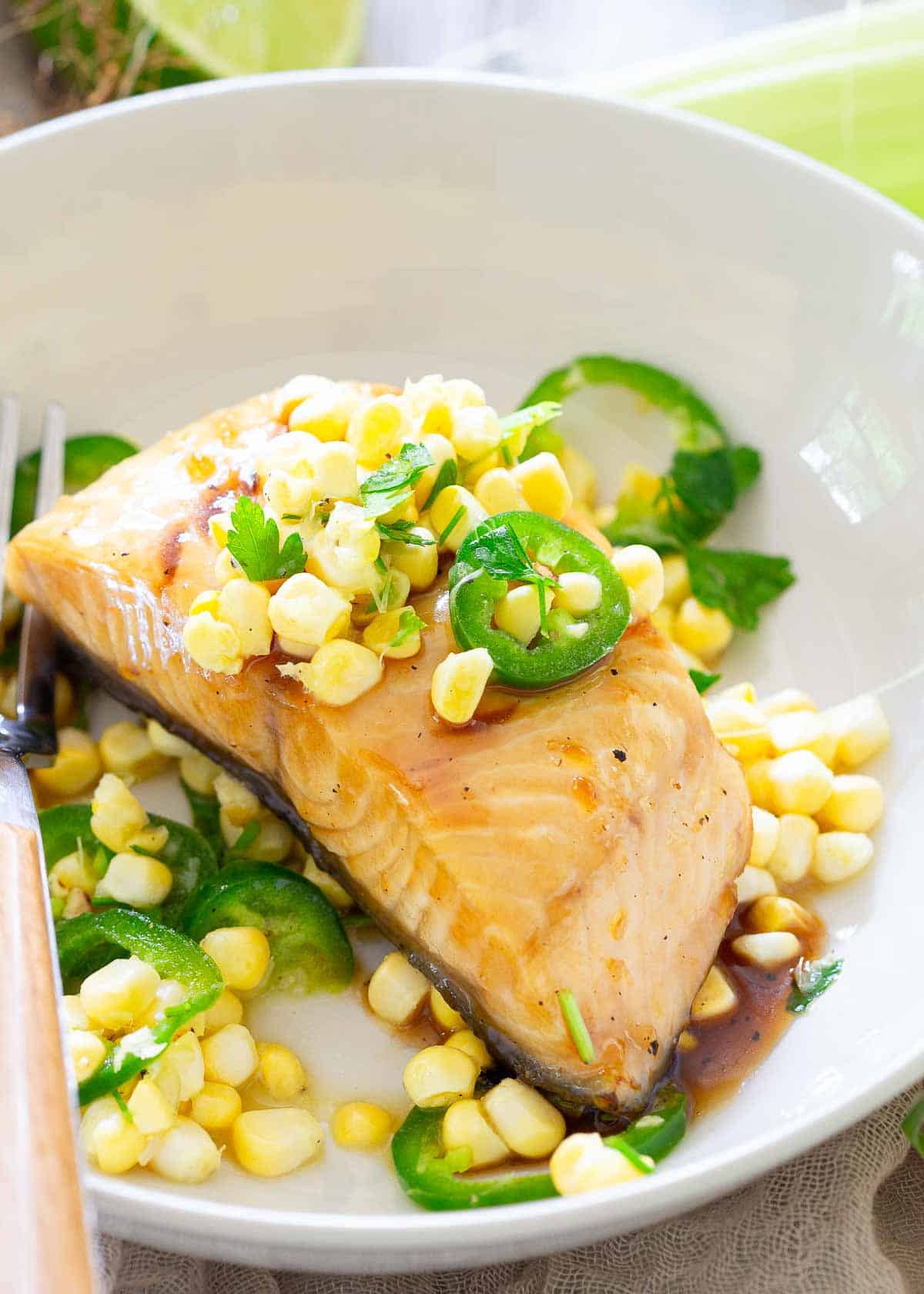 A plate of cooked sablefish topped with corn kernels and sliced jalapeños, garnished with parsley. A fork rests on the edge of the plate.