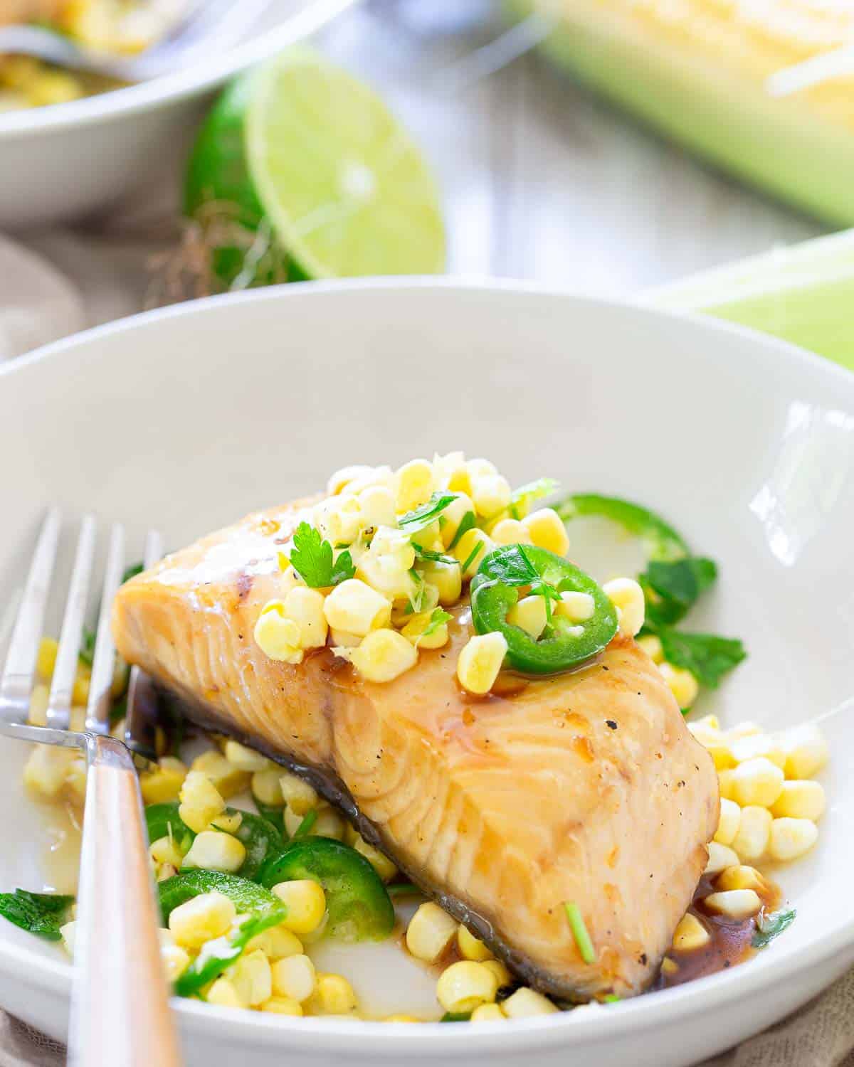 A piece of cooked black cod topped with corn kernels, sliced jalapeños, and herbs, served on a white plate with a fork.