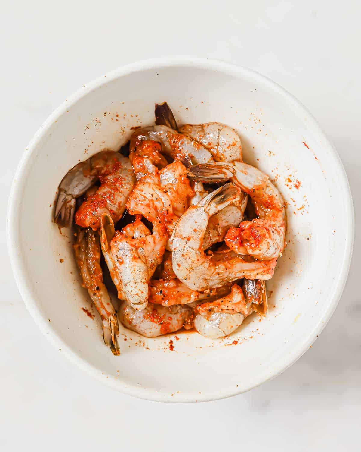 A white bowl filled with raw shrimp seasoned with spices.