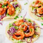 Three shrimp tostadas garnished with purple cabbage, green onions, and herbs on a white surface.