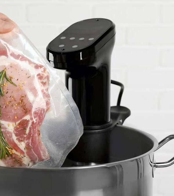 A hand lowering a vacuum-sealed bag of raw meat with herbs into a pot with an immersion circulator for sous vide cooking.