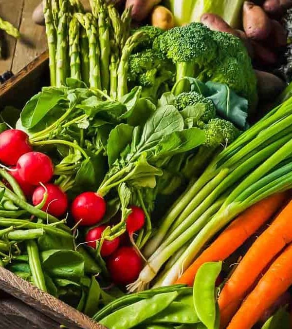 A variety of fresh vegetables including carrots, radishes, asparagus, and greens in a wooden crate on a rustic table.