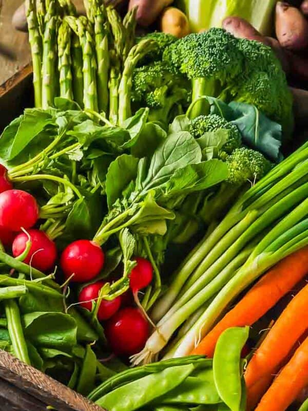 A variety of fresh vegetables including carrots, radishes, asparagus, and greens in a wooden crate on a rustic table.