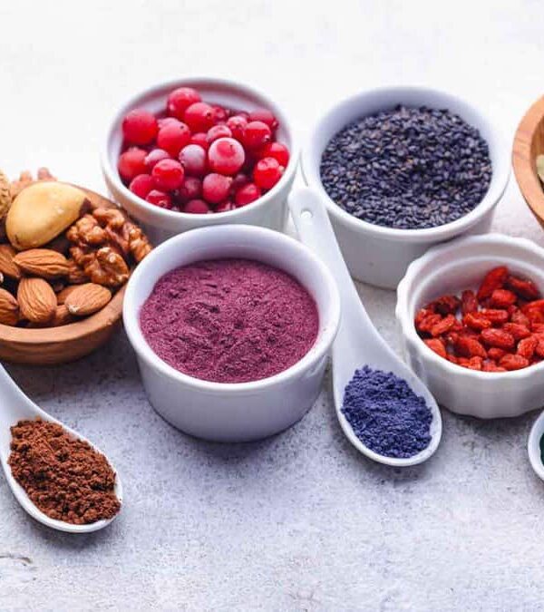 A variety of small bowls and spoons containing different seeds, nuts, and powdered spices arranged on a light surface.