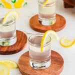 Three shot glasses filled with clear liquid, each garnished with a lemon slice, are placed on wooden coasters. Lemon slices are scattered around on a white surface.