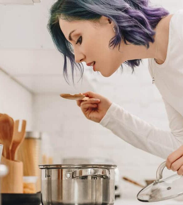 A woman with blue and purple hair tasting food from a pot in a kitchen, holding a wooden spoon and lifting a pot lid.
