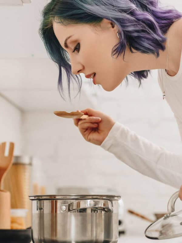 A woman with blue and purple hair tasting food from a pot in a kitchen, holding a wooden spoon and lifting a pot lid.