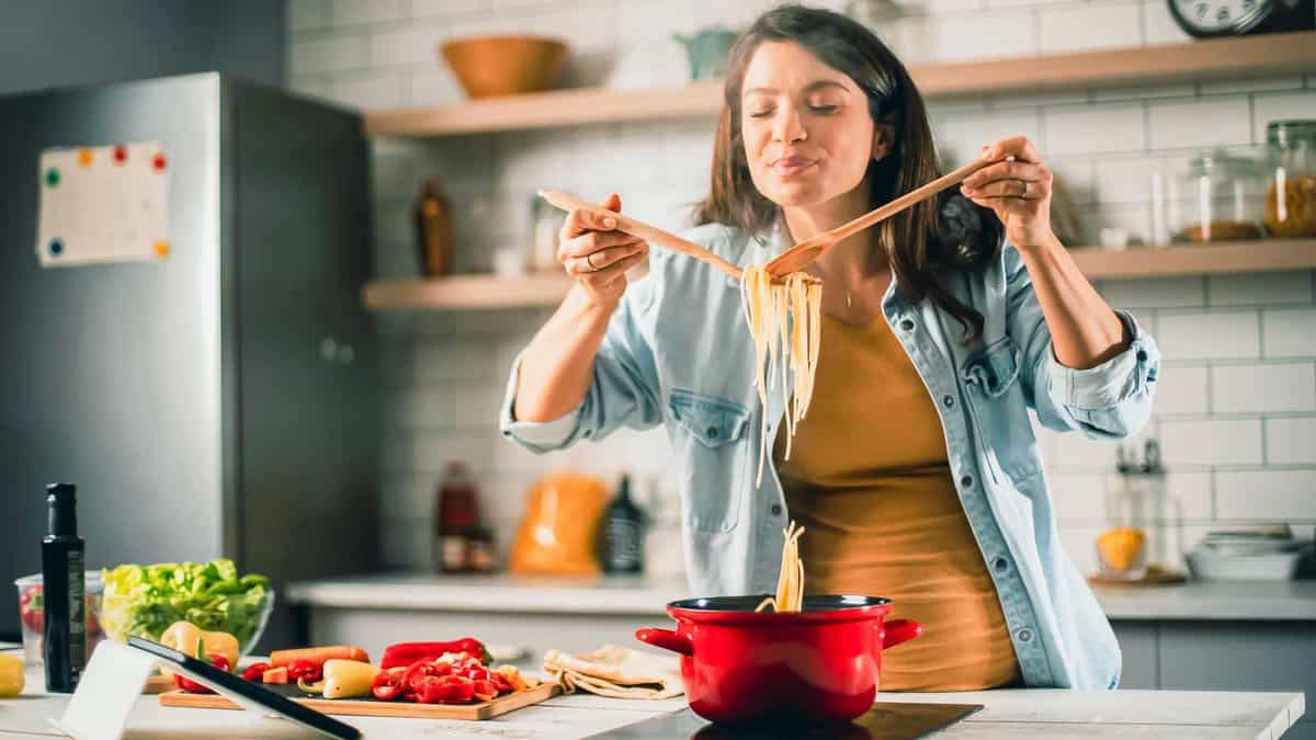 A woman cooking in a kitchen, lifting spaghetti from a red pot using wooden utensils. Various ingredients are visible on the counter.