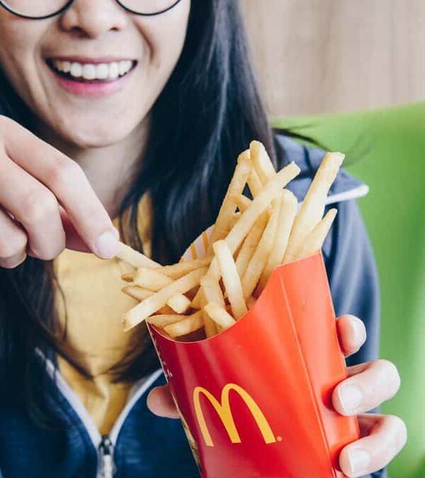 Woman in a yellow shirt, smiling, picking a French fry from a McDonald's container at a restaurant table.