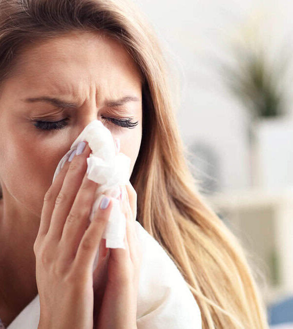 A woman with long hair blowing her nose into a tissue, appearing to be sick or having an allergic reaction.