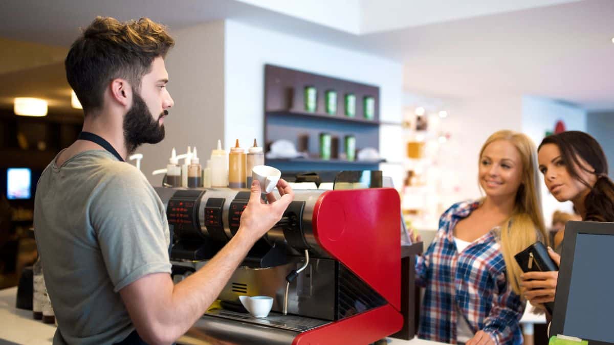 A barista explaining coffee options to two female customers at a cafe counter.