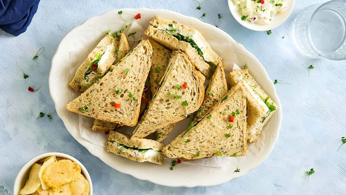 A plate of triangular sandwiches with various fillings, garnished with herbs and diced red peppers. A side dish of chips and a small bowl of additional filling are also visible.