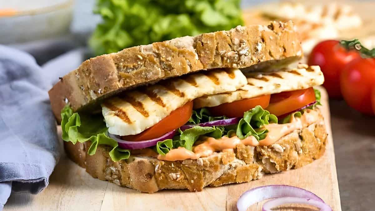 A grilled cheese sandwich on whole grain bread with slices of tomato, lettuce, red onion, and a creamy spread on a wooden surface, surrounded by leafy greens and cherry tomatoes.