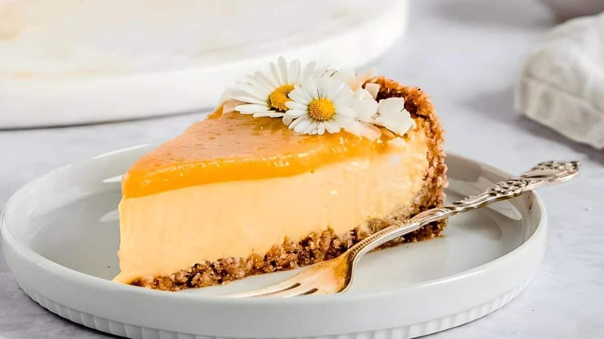 A slice of orange cheesecake garnished with white flowers, served on a white plate with a silver fork.
