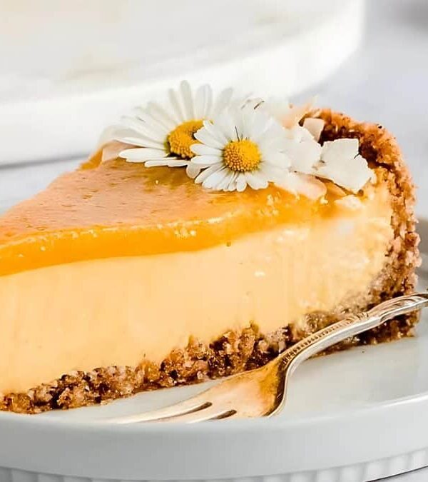 A slice of orange cheesecake garnished with white flowers, served on a white plate with a silver fork.