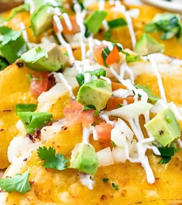 A close-up of three stuffed and folded yellow corn tacos topped with diced avocado, tomato, onion, and drizzled with white sauce.