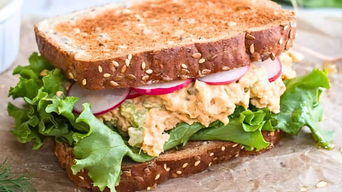 A close-up of a sandwich made with whole grain bread, lettuce, sliced radishes, and a creamy filling, placed on a piece of parchment paper.