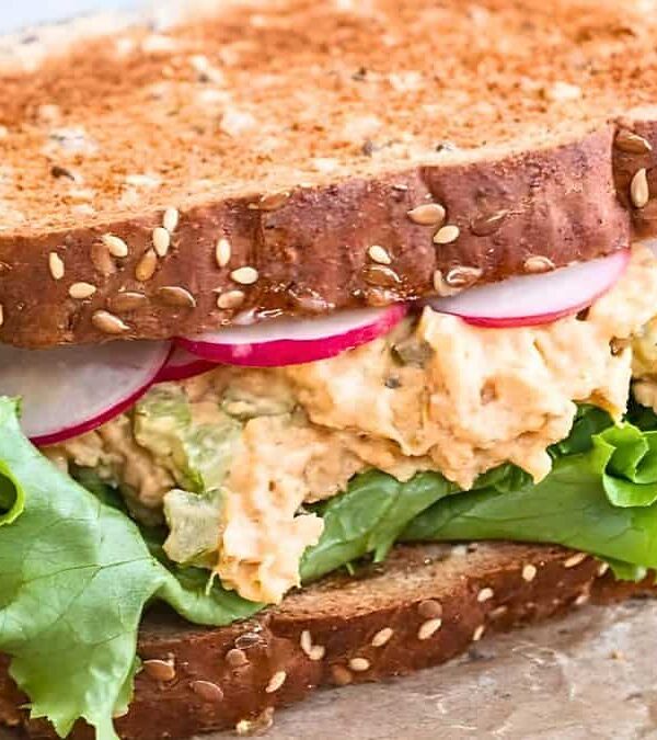 A close-up of a sandwich made with whole grain bread, lettuce, sliced radishes, and a creamy filling, placed on a piece of parchment paper.